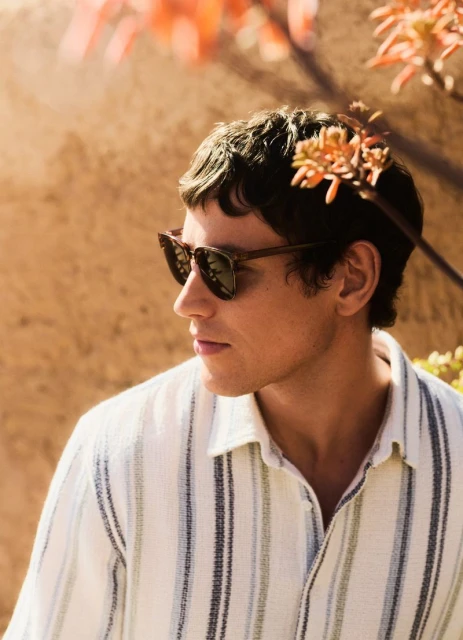 Outdoor portrait of a young man with dark hair wearing sunglasses and a striped shirt, with a textured wall and flowers in the background.