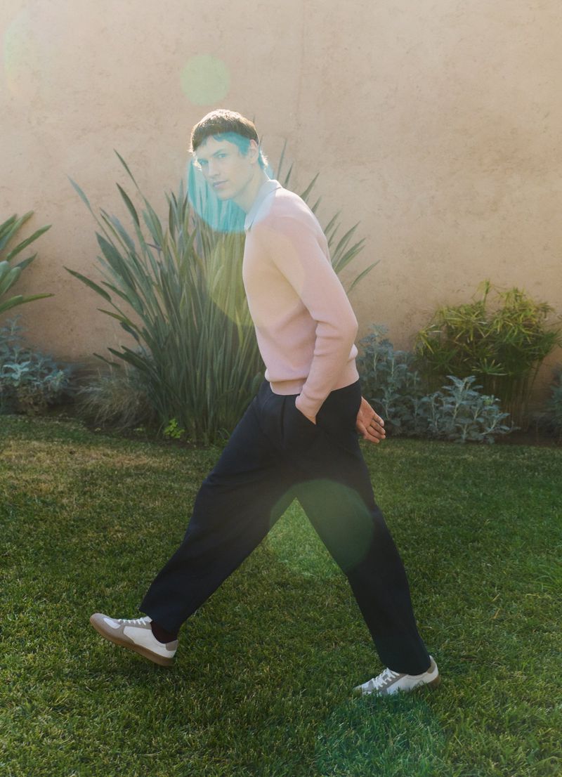 Young man poses in an outdoor shot, showcasing casual fashion with a pink sweater, dark pants, and white sneakers on a green lawn.