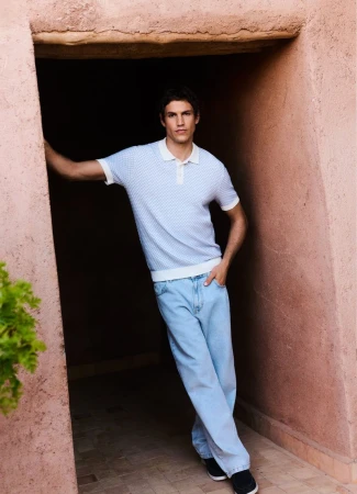 A young adult male model poses against a peach-colored wall in a doorway wearing a short-sleeved knit shirt and light-wash denim jeans.