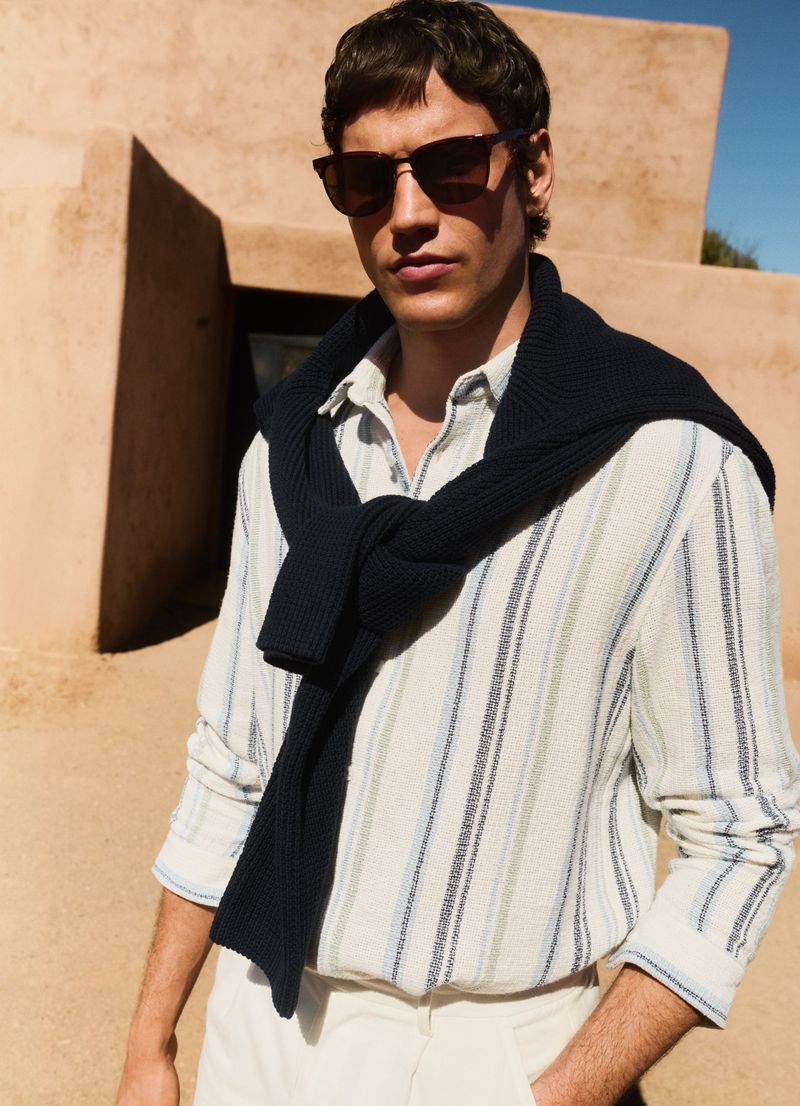 A man in sunglasses and a striped shirt poses outdoors with a dark sweater draped over his shoulders, looking towards the viewer.