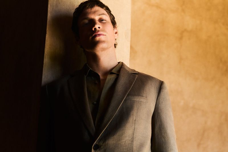 Close-up studio shot of an adult male model in a tan suit and shirt leaning against an orange wall with soft, diffused natural lighting.