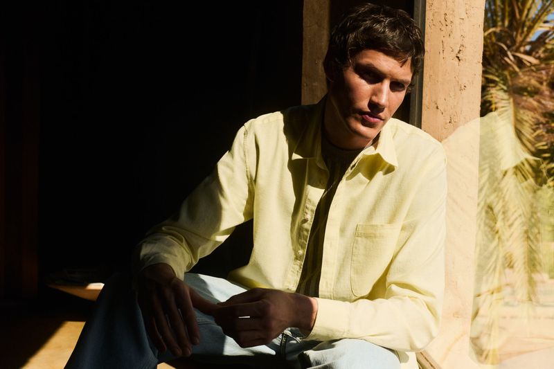 Portrait of a young man wearing a yellow button-down shirt and jeans, seated indoors with natural light creating strong shadows.