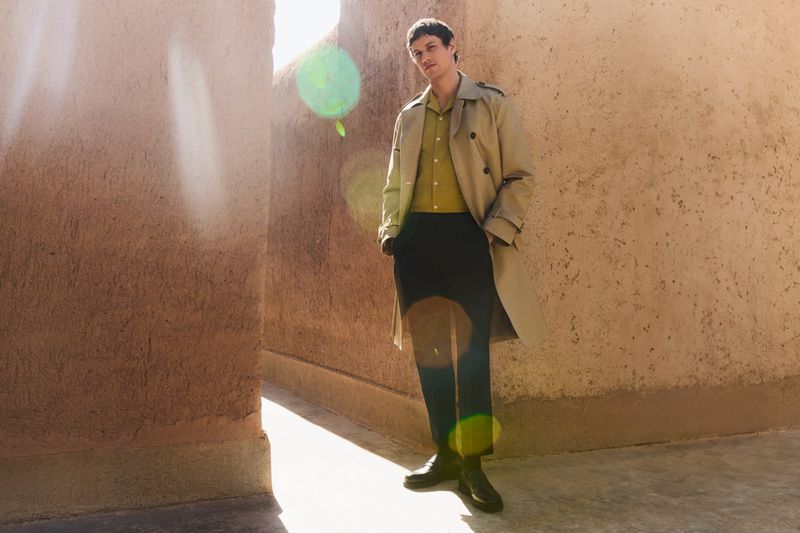 A young adult male poses in a tan trench coat and dark pants in a sunlit alleyway, blending fashion with an urban environment.