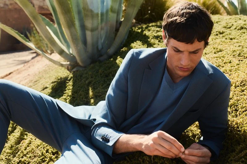 A handsome man lies in a suit on a green grassy area, staring down at his fingers, with a large agave plant in the background.