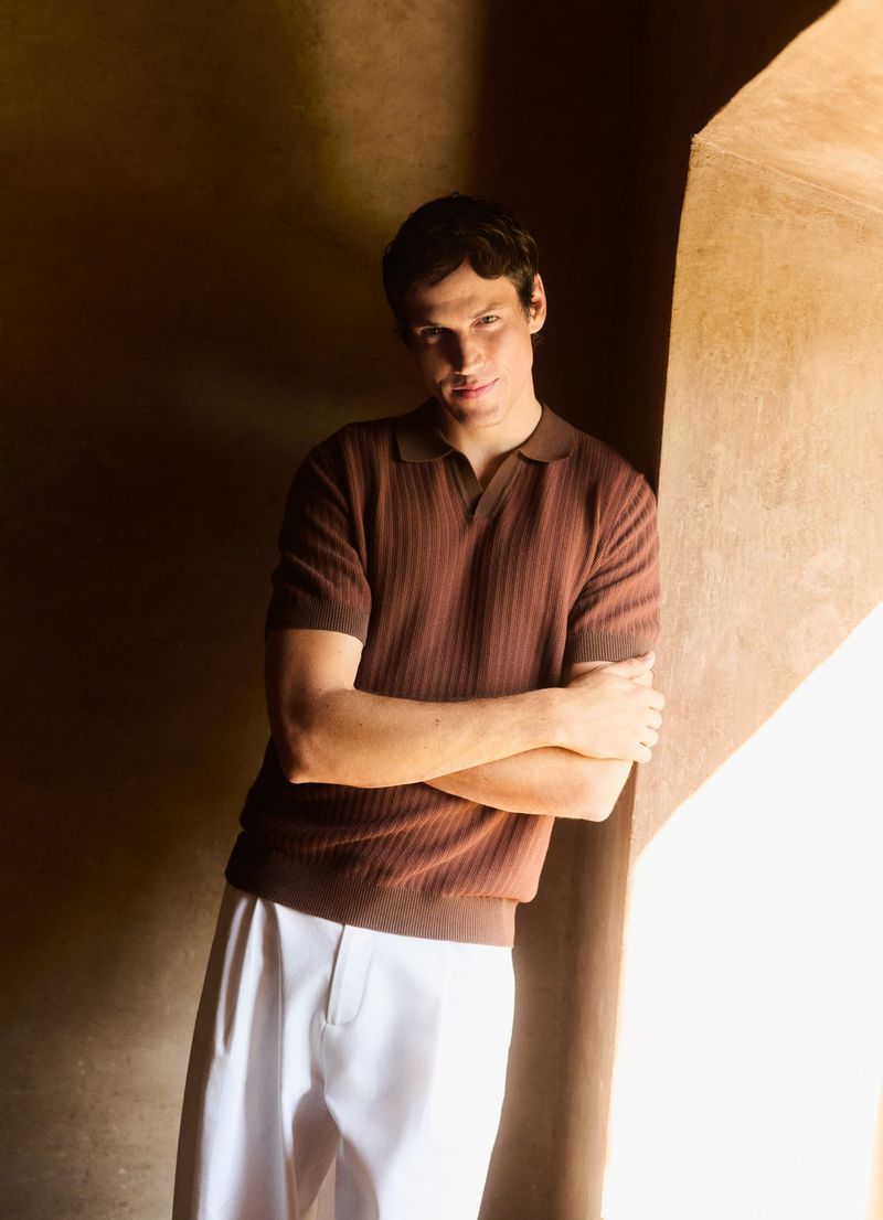A young adult male in a brown polo shirt and white pants leans against a textured wall in a naturally lit indoor setting.