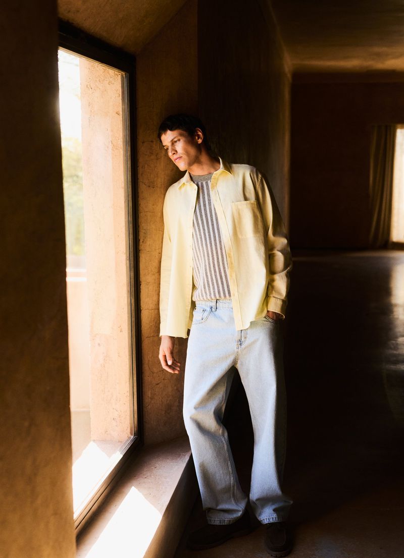 Adult male in light-wash jeans, striped shirt, and a pale yellow button-down shirt looking out a window in a sunlit, ochre-toned room.
