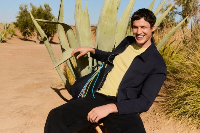 Smiling man in jacket and yellow tee shirt poses in a desert landscape with agave plants under a clear sky on a sunny day.