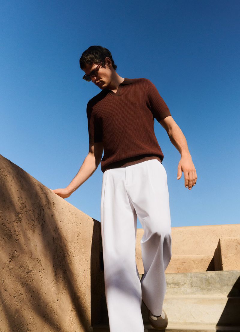 Man in brown shirt and white pants walks down outdoor stairs on a sunny day against a clear blue sky, wearing sunglasses.