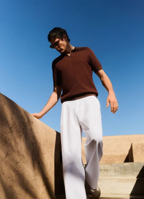 Man in brown shirt and white pants walks down outdoor stairs on a sunny day against a clear blue sky, wearing sunglasses.