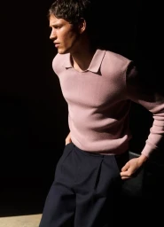 Male model wearing a pink polo shirt and dark trousers posed against a dark background with strong directional lighting.