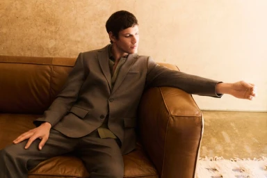 Model wearing a brown suit jacket seated on a brown leather couch in a warm-toned interior