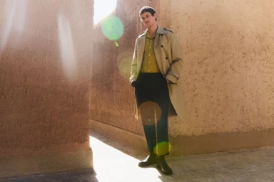 Male model wearing a beige trench coat and dark trousers standing between textured earthen walls.