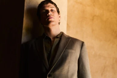 Model wearing a dark blazer leaning against a warm textured wall in a low-light portrait