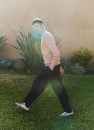 Model wearing a pink sweater and dark trousers walking across a grassy lawn with plants against a warm wall