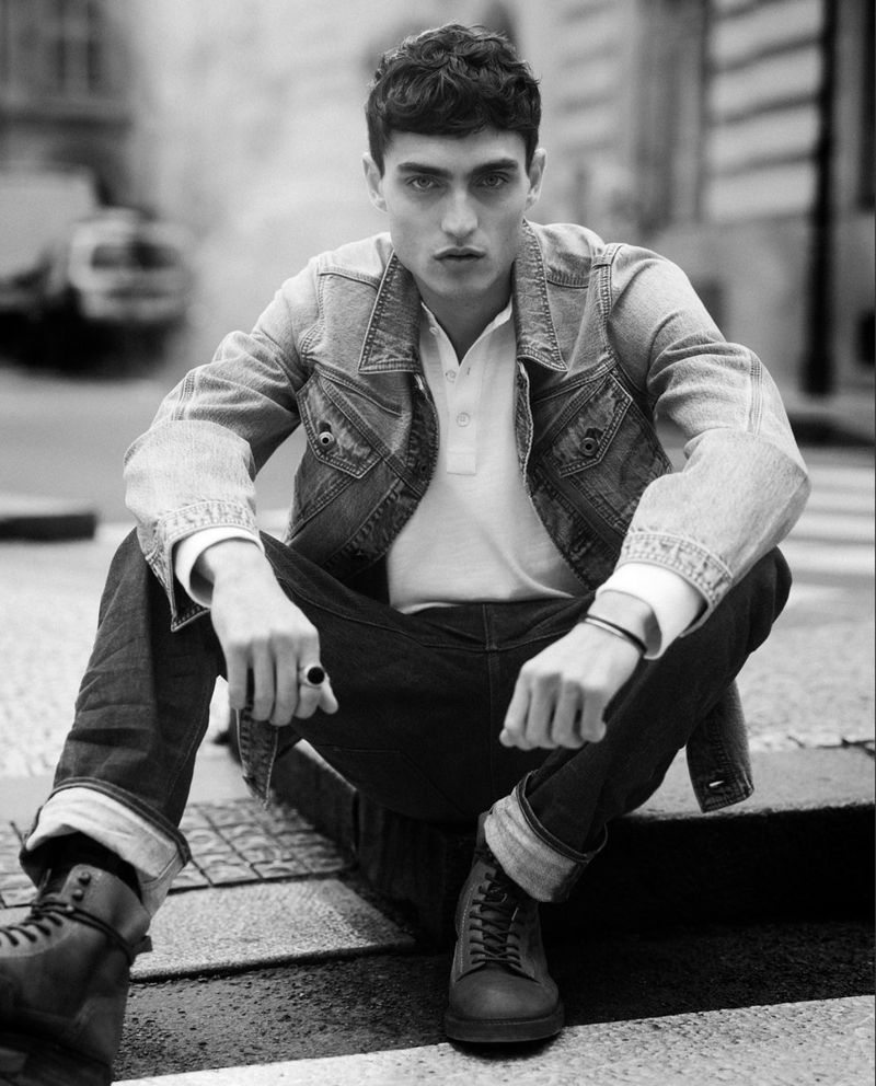 Black and white portrait of a dark-haired male model in a denim jacket sitting in an urban environment looking directly at the camera.