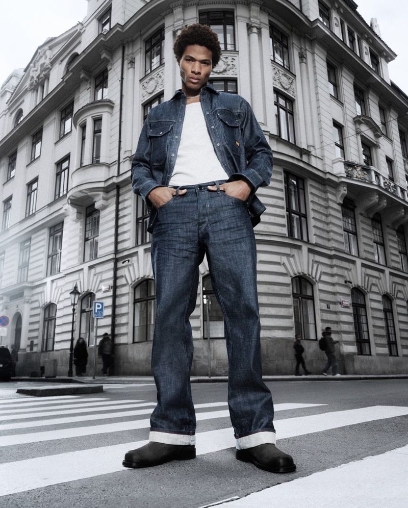 A young adult man stands in a crosswalk wearing a denim jacket and jeans, in front of a grey stone building in an urban setting.
