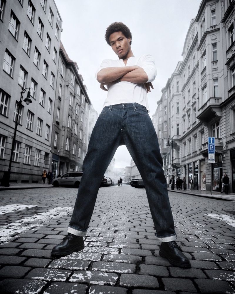 Low-angle shot of a young man with folded arms standing on a cobblestone street framed by tall city buildings.
