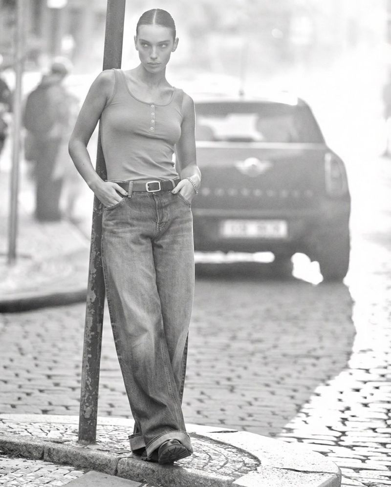 A young adult woman in denim leans on a pole in a black and white outdoor fashion shot with a car in the blurred background.