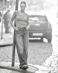 Monochrome fashion campaign image of a model leaning against a pole on a cobblestone street with a car parked in the background.