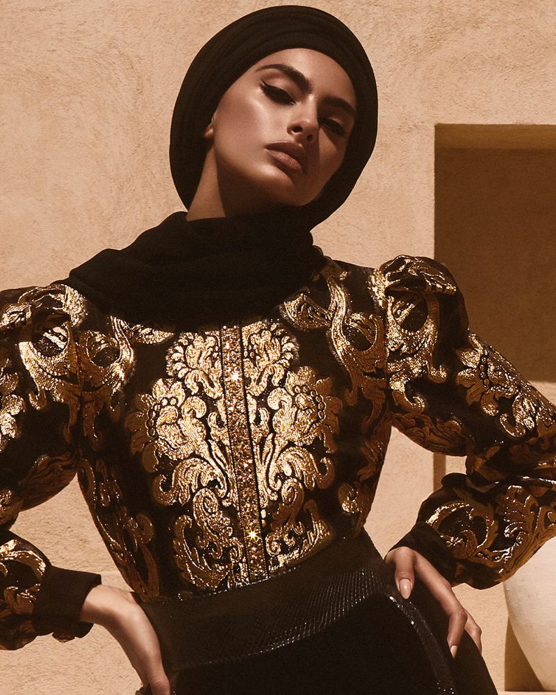 Portrait of a woman with dark hair wearing a black turban and a gold embroidered jacket, posing in front of a textured wall.