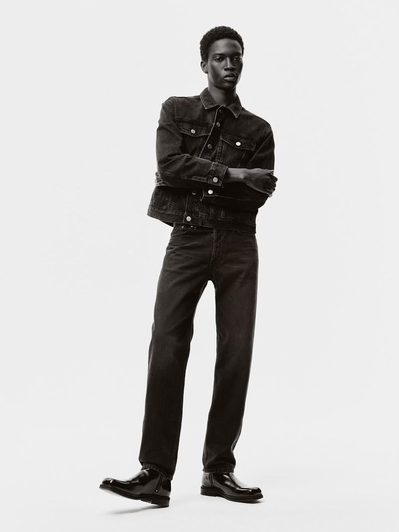 A male fashion model poses in a denim jacket and jeans in a monochrome studio shot with arms crossed against a white backdrop.