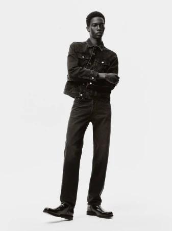 A male fashion model poses in a denim jacket and jeans in a monochrome studio shot with arms crossed against a white backdrop.