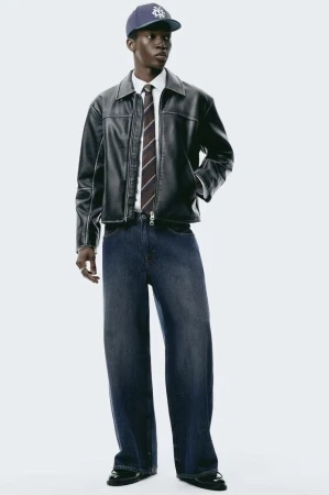 Male fashion model poses in a black leather jacket, white collared shirt, striped tie, wide-leg jeans, cap, and black loafers indoors.