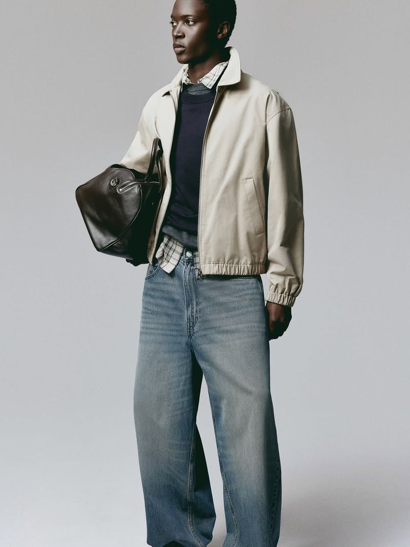 A Black male model stands against a plain grey background, wearing a cream bomber jacket, navy sweater, plaid shirt, and wide-leg denim jeans.