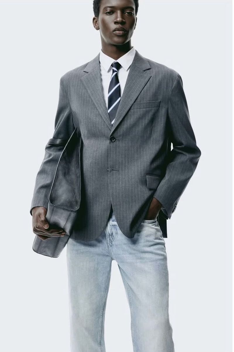 Male fashion model wears a grey pinstripe suit with a white shirt and blue tie against a bright white background in a studio shot.