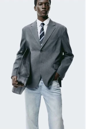 Male fashion model wears a grey pinstripe suit with a white shirt and blue tie against a bright white background in a studio shot.