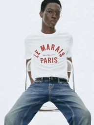 Model Limamu Mbaye wearing a white graphic t-shirt and blue jeans seated on a clear chair against a white background.
