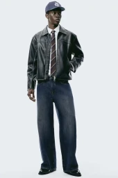 Model Limamu Mbaye wearing a black faux leather jacket and dark denim jeans against a plain white background.
