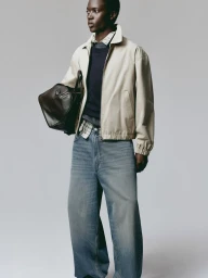 Model wearing a light jacket, dark sweater, and denim jeans holding a dark leather duffel bag against a light grey background.