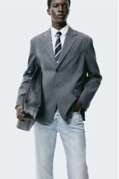 Model wearing a grey pinstripe blazer, white shirt, striped tie, and light wash jeans holding a dark bag against a plain white background