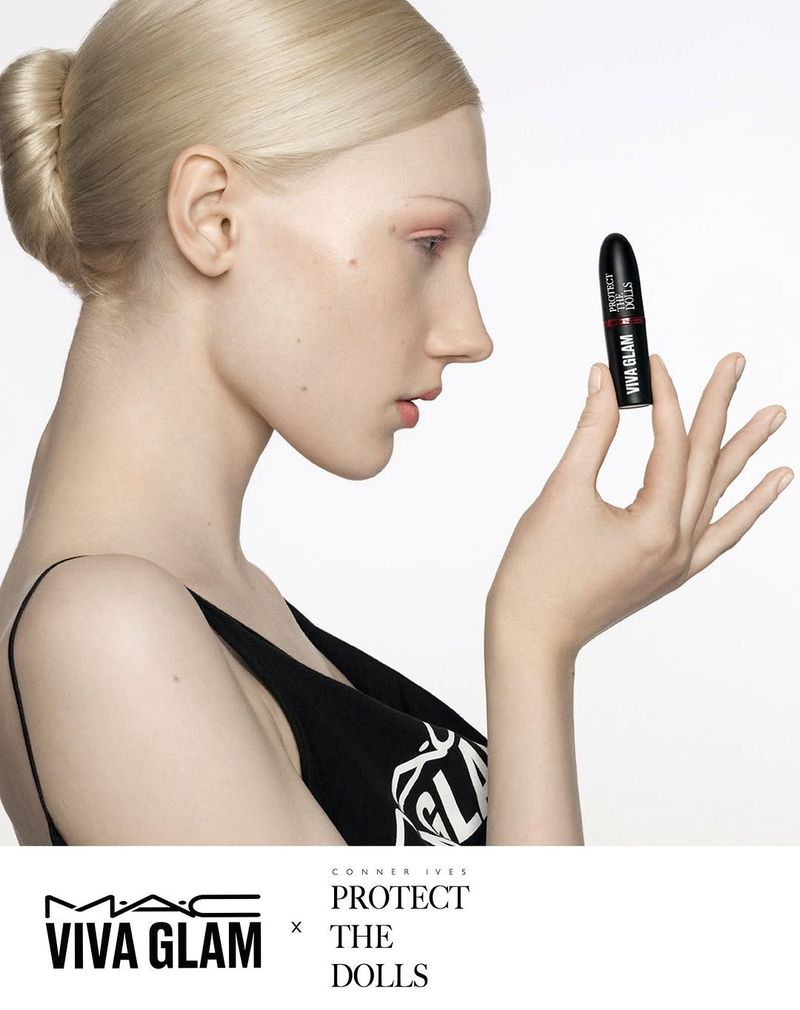 Woman poses with lipstick for MAC Viva Glam campaign, featuring a light skin tone, blonde hair, black top, and a light studio backdrop.