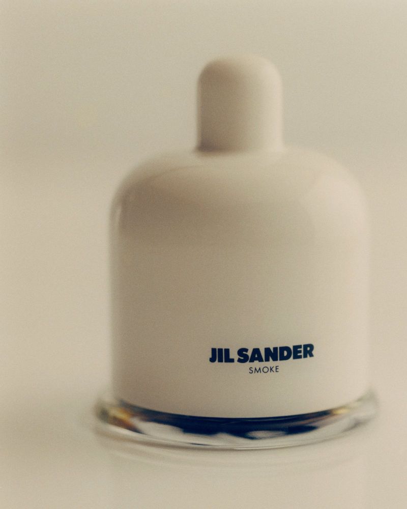 Studio shot of a white ceramic perfume bottle with a clear glass base, featuring the Jil Sander brand and 'Smoke' fragrance name.