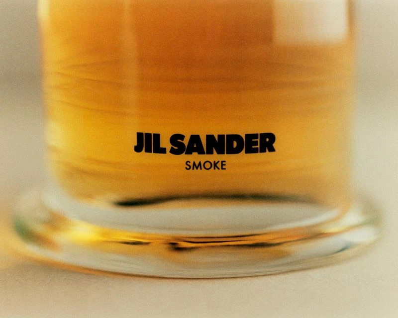 Close-up of a clear glass container filled with amber-colored liquid, featuring the Jil Sander Smoke logo in black letters.
