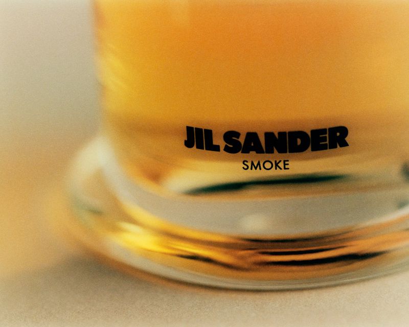 Jil Sander 'Smoke' advert features a close-up of a translucent perfume bottle with black sans-serif text against a neutral background.