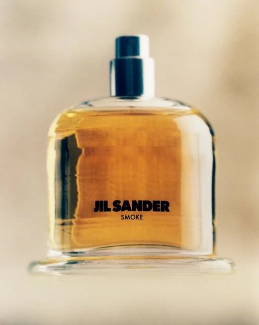 Studio shot of Jil Sander Smoke perfume bottle, amber liquid, clear glass, silver cap on a reflective surface, cream gradient background.