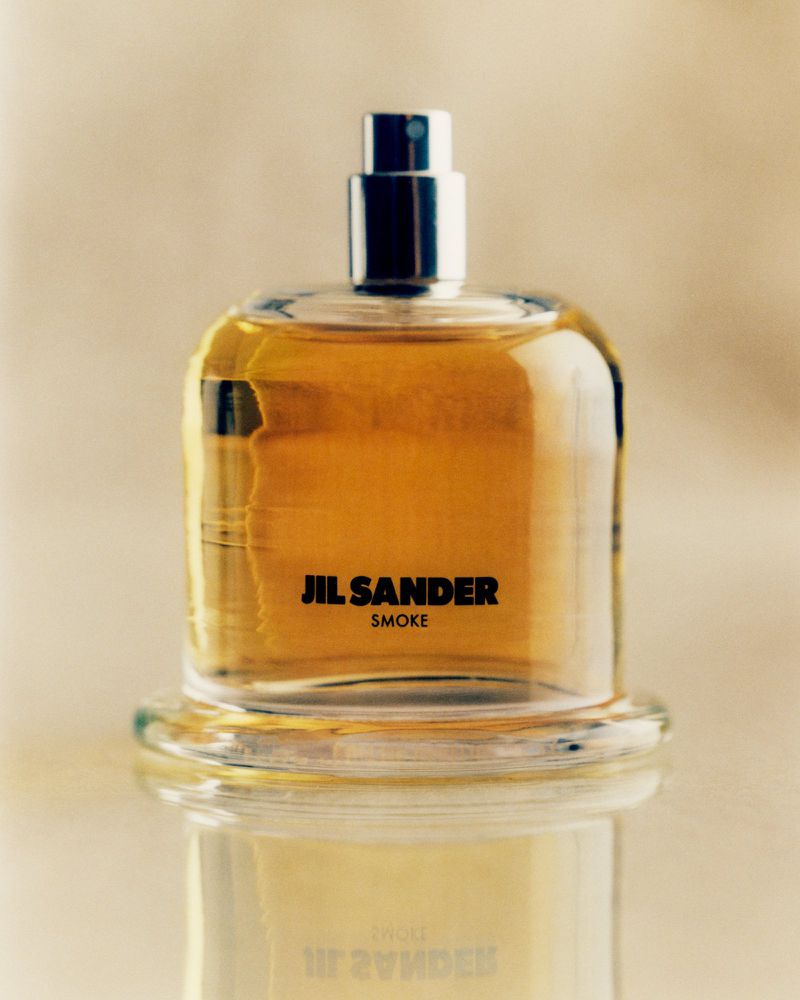 Jil Sander Smoke perfume bottle on a glass surface, featuring a clear square bottle, silver nozzle, and warm amber-toned liquid.