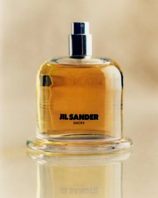 Jil Sander Smoke perfume bottle on a glass surface, featuring a clear square bottle, silver nozzle, and warm amber-toned liquid.
