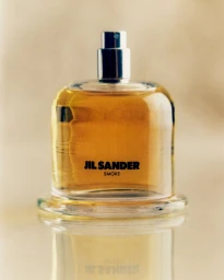 Glass Jil Sander Smoke perfume bottle containing amber liquid resting on a reflective surface against a neutral background.