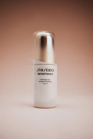 A white Shiseido Benefiance serum bottle with a silver cap on a peach-colored background, featuring product branding and marketing text.