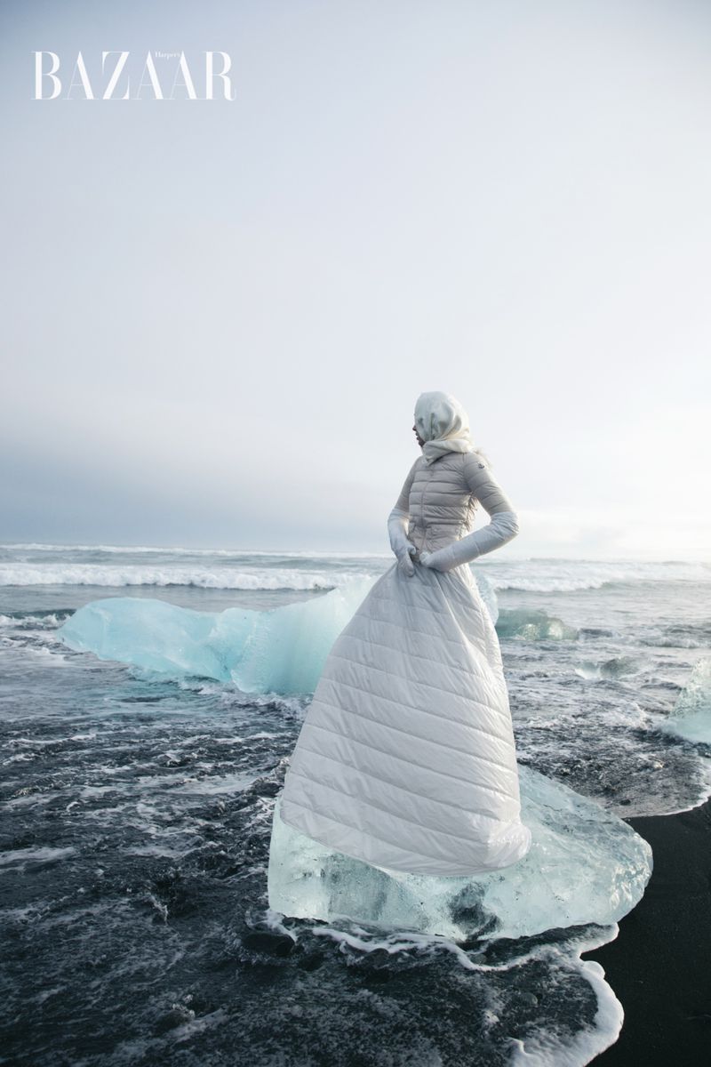 Fashion shot of a woman wearing a long white quilted dress and jacket, standing on an iceberg in a dark, wavy ocean in Iceland.