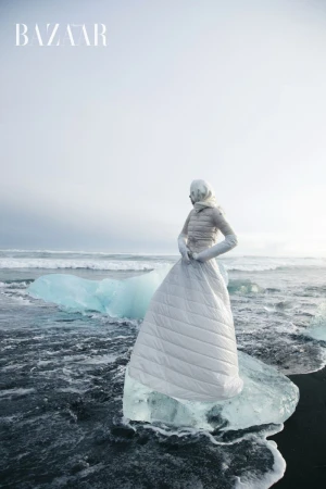 Fashion shot of a woman wearing a long white quilted dress and jacket, standing on an iceberg in a dark, wavy ocean in Iceland.