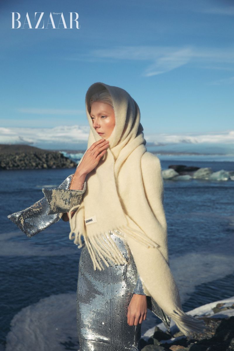 Fashion shot of a woman in a silver sequin dress and cream scarf, posing on a rocky shore with a glacial lake and mountains in the background.