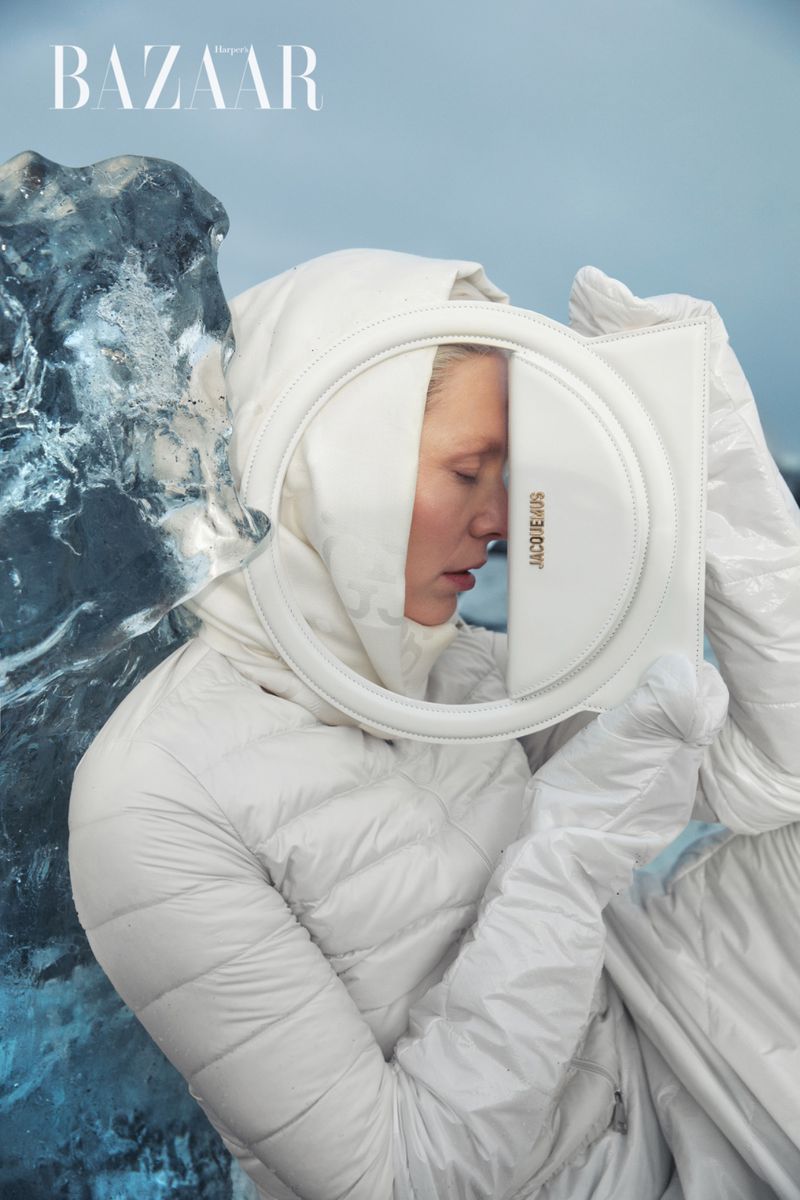 Fashion editorial shot of a blond woman in a puffy white coat and gloves, holding a round white purse, next to a block of ice.