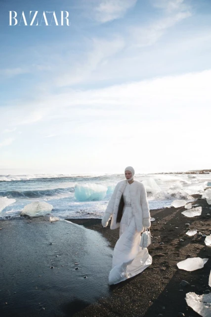 Fashion shoot on a black sand beach featuring a model in a white gown, accessorized with a jacket and small handbag, waves and ice.