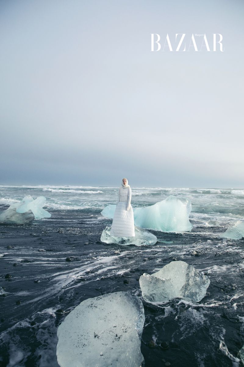 Fashion editorial image featuring a model in a white dress posing on a black sand beach surrounded by ice chunks near the ocean.