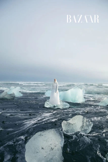 Fashion editorial image featuring a model in a white dress posing on a black sand beach surrounded by ice chunks near the ocean.